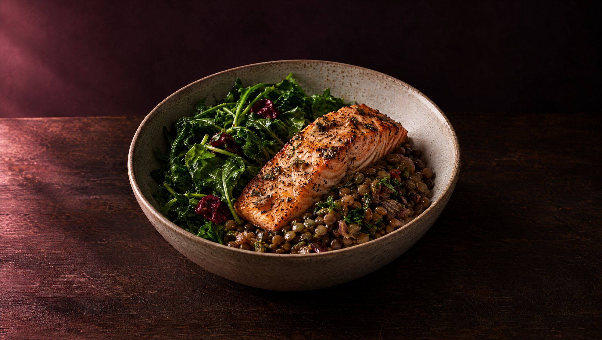 A ceramic bowl of leafy greens, lentils and grilled salmon on a dark wooden table in soft rose side light.