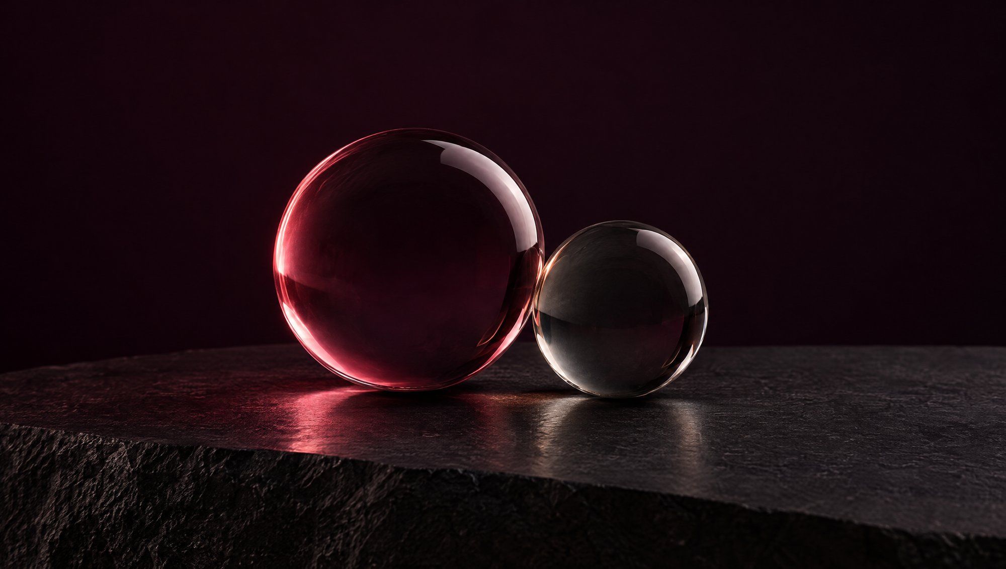 Two clear glass spheres of different sizes touching on dark stone, the larger one catching rose light.